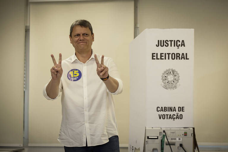 Fotografia mostra o governador Tarcísio de Freitas em uma seção de votação. Ele é um homem branco, usa camisa branca e adesivo com "15" no peito e faz sinal de "V" com os dedos