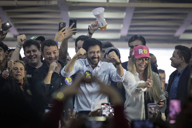 Fotografia mostra o o prefeito reeleito Ricardo Nunes (MDB) comemorando a vitória nas eleições de 20224. Há também um grupo de pessoas ao redor dele, com várias pessoas sorrindo e levantando os braços em sinal de celebração. No centro, Nunes usa camisa branca e uma mulher com um boné rosa estão sorrindo e fazendo gestos de vitória. Ao fundo, há várias pessoas segurando celulares e outros objetos, todos parecendo animados