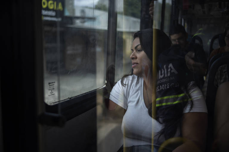 A imagem mostra uma mulher sentada em um ônibus, olhando pela janela. Ela está vestindo uma camiseta branca e parece pensativa. Ao fundo, é possível ver outras pessoas sentadas e a parte interna do ônibus, com reflexos na janela.