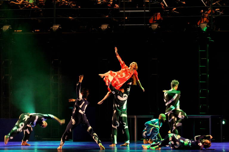 A imagem mostra uma apresentação de dança em um palco. No centro, uma dançarina com um vestido vermelho com bolinhas brancas é levantada por um dançarino. Outros dançarinos, vestidos com trajes pretos e brancos, estão em várias poses ao redor. Ao fundo, uma orquestra é visível, com músicos tocando instrumentos. A iluminação é dramática, com luzes verdes e azuis no palco.