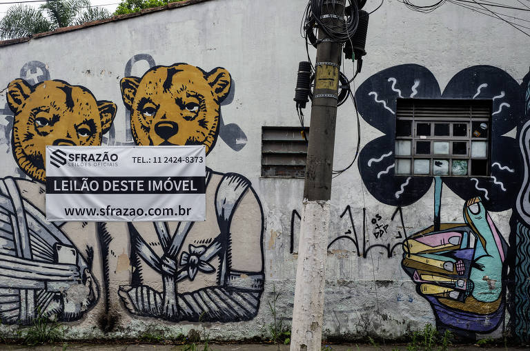 A imagem mostra um mural de grafite em uma parede. O mural apresenta duas figuras com cabeças de urso, uma delas vestindo uma blusa e a outra com um vestido. Ao lado, há um anúncio em uma placa branca que diz 'LEILÃO DESTE IMÓVEL' e inclui informações de contato e um site. A parede também possui uma janela com grades e um desenho de uma mão segurando um copo.