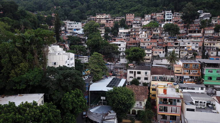 A imagem mostra uma visão aérea de uma comunidade localizada em uma área montanhosa e arborizada. As casas são predominantemente de alvenaria e variam em tamanhos e cores, indicando uma construção densa e vertical em algumas áreas. No centro da imagem, um prédio de telhado branco destaca-se por ter painéis solares instalados, o que sugere um projeto de energia renovável na região. A paisagem ao redor é marcada por muita vegetação, com árvores altas contrastando com as construções urbanas.