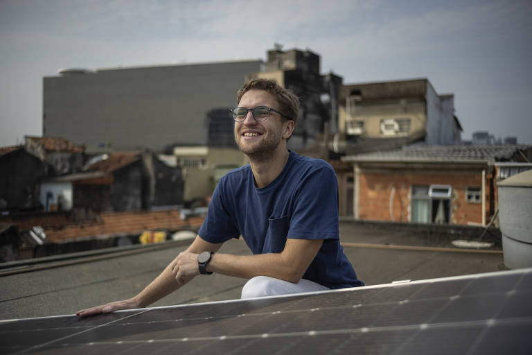 A imagem mostra um homem sorrindo enquanto está ajoelhado ao lado de um painel solar, em um telhado de uma área urbana. Ele está usando uma camiseta azul e óculos, transmitindo uma expressão de satisfação ou entusiasmo. Ao fundo, há prédios residenciais e comerciais, indicando um ambiente urbano denso. A luz do dia é suave, sugerindo um momento de manhã ou fim de tarde.