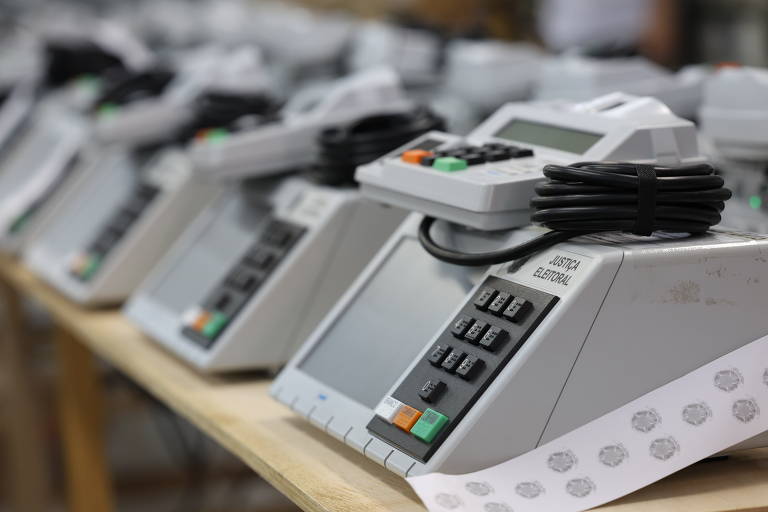 A imagem mostra uma linha de máquinas de votação eletrônicas dispostas em uma mesa. Cada máquina possui um visor e um teclado com botões coloridos, incluindo verde, laranja e preto. Os cabos estão conectados a cada unidade, e há uma folha com selos ou marcas na parte inferior da imagem.