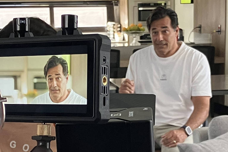 A imagem mostra um homem sentado em um sofá em um ambiente de estúdio, sendo filmado por uma câmera. O homem está usando uma camiseta branca e parece estar falando ou explicando algo. Na tela da câmera, ele é visto em close, com uma expressão facial que sugere que ele está se comunicando de forma séria. Ao fundo, há uma cozinha moderna com uma mesa e cadeiras, além de uma fruteira. A câmera está montada em um tripé e há um braço de suporte visível.