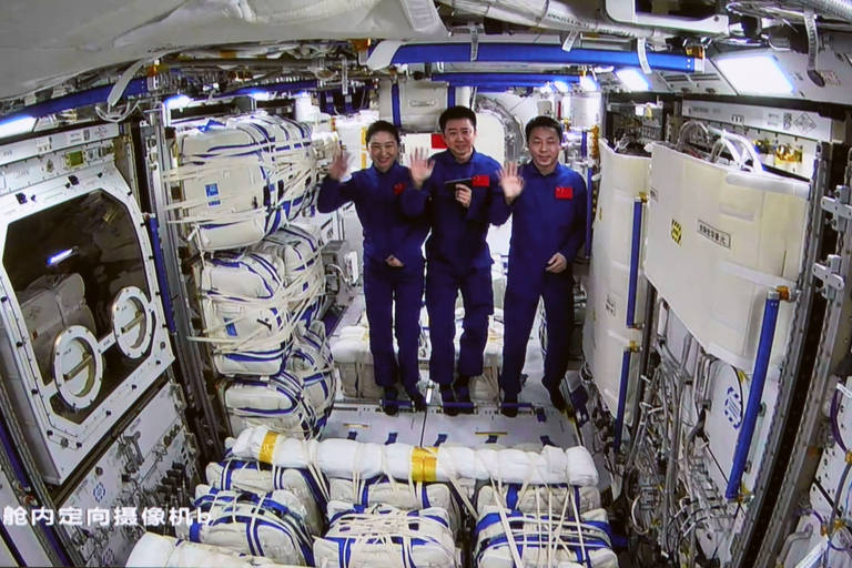 Três astronautas estão dentro de uma estação espacial, vestindo trajes azuis. Eles estão sorrindo e acenando para a câmera. O interior da estação é equipado com painéis e equipamentos, e há vários pacotes e cabos ao fundo.