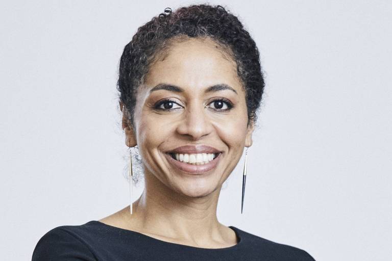 A imagem mostra uma mulher com cabelo cacheado e escuro， usando uma blusa preta. Ela está sorrindo e com os braços cruzados， em um fundo claro. A mulher usa brincos longos e finos.