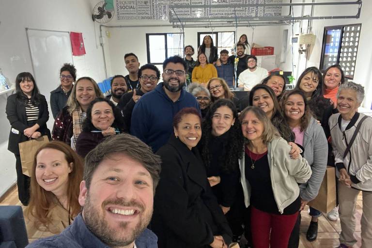 Uma foto em grupo de várias pessoas em um ambiente interno. Algumas pessoas estão em pé ao fundo， enquanto outras estão mais próximas da câmera. O ambiente possui paredes brancas e uma lousa. Há também prateleiras com objetos e plantas. As pessoas estão sorrindo e parecem estar se divertindo.