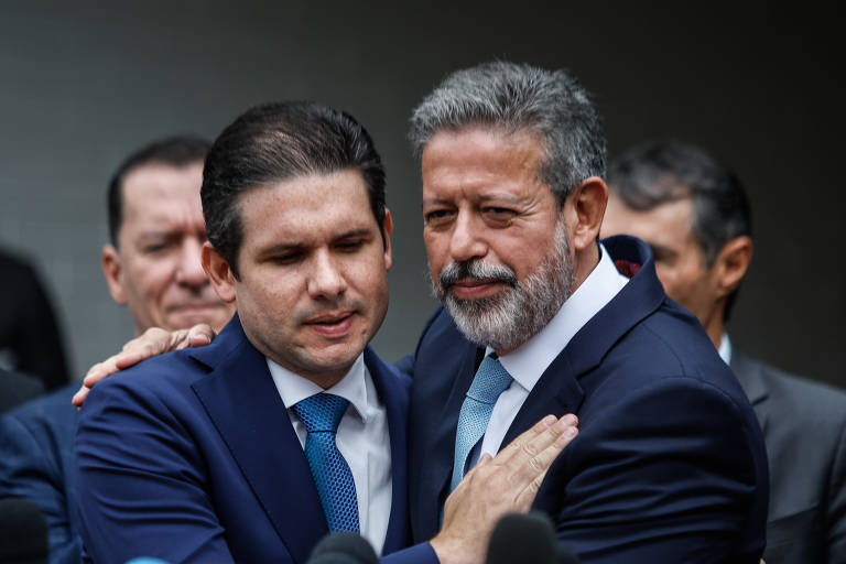 A imagem mostra dois homens em trajes formais se abraçando. O homem à esquerda tem cabelo escuro e liso, e usa um terno azul com uma gravata clara. O homem à direita tem cabelo grisalho e uma barba bem cuidada, vestindo um terno escuro e uma gravata azul. Ao fundo, outras pessoas estão visíveis, mas não são o foco da imagem.