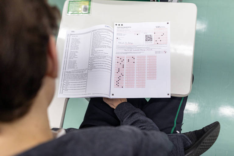 Um aluno está sentado em uma mesa， segurando uma folha de prova. A folha contém uma lista de itens e um gráfico com quadrados preenchidos em vermelho. O aluno é visto de costas， e a mesa é de cor clara. O chão é de um tom verde.