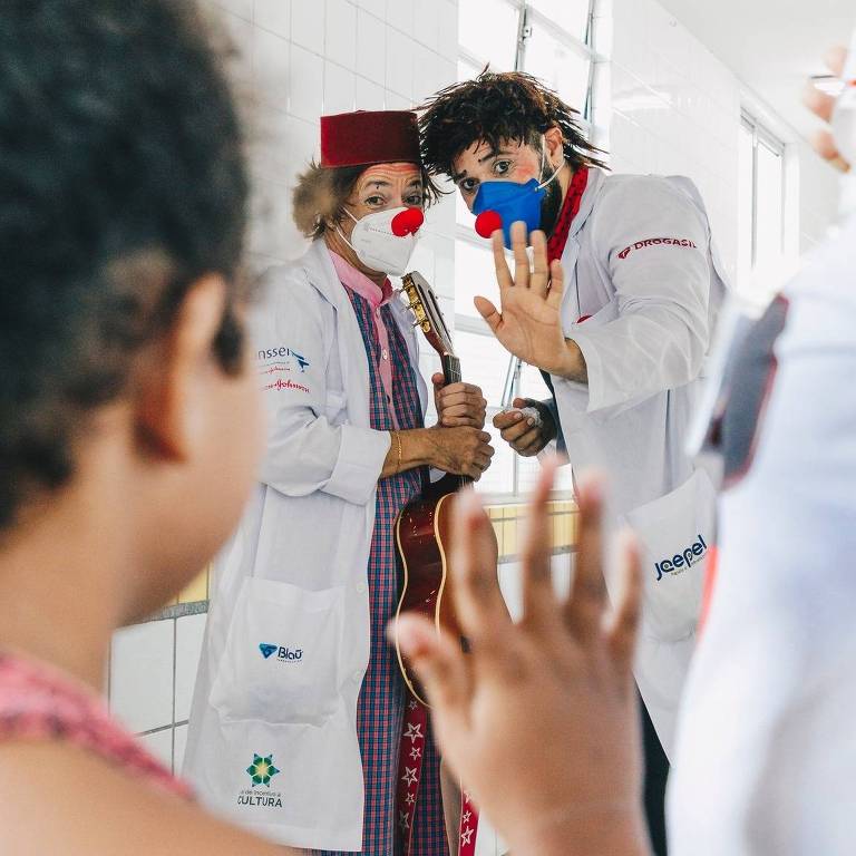 Uma cena em um hospital onde dois palhaços, um segurando um violão, estão interagindo com uma criança. Os palhaços estão usando máscaras e fantasias coloridas, com um palhaço usando um fez vermelho e o outro uma máscara azul com um nariz vermelho. A criança, vista de trás, está acenando para eles.