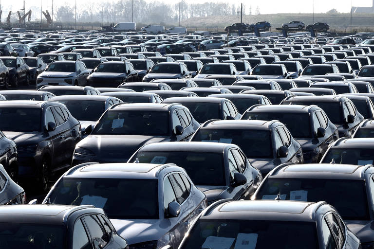 A imagem mostra um grande estacionamento com várias fileiras de carros, predominantemente de cor prata. Os veículos estão organizados em linhas, com muitos deles visíveis ao fundo. O ambiente parece ser ao ar livre, com um céu claro e algumas árvores ou estruturas ao longe.