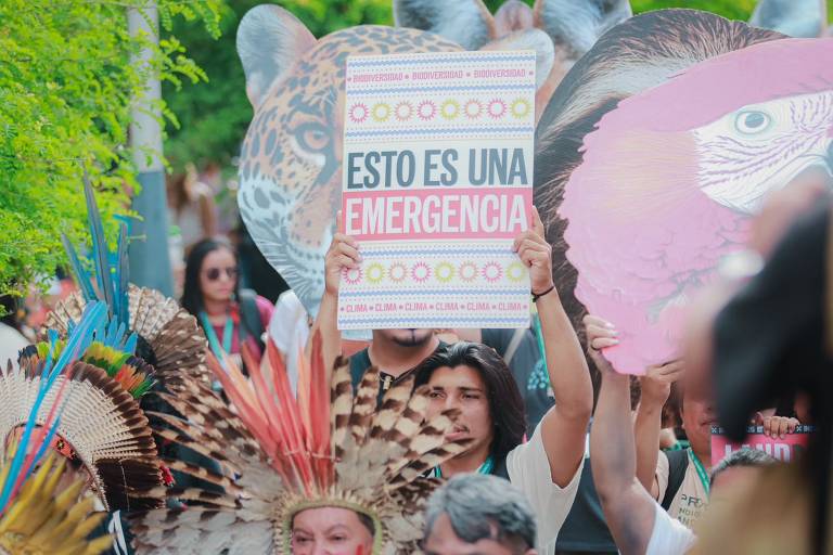 A imagem mostra um grupo de pessoas participando de um protesto. Um homem em destaque segura um cartaz que diz 'ESTO ES UNA EMERGENCIA' em letras grandes e coloridas. Ao fundo, há pessoas com trajes tradicionais e adereços, incluindo penas. Também são visíveis cartazes em forma de animais, como um jaguar e um pássaro, que representam a biodiversidade. A cena é cercada por vegetação.