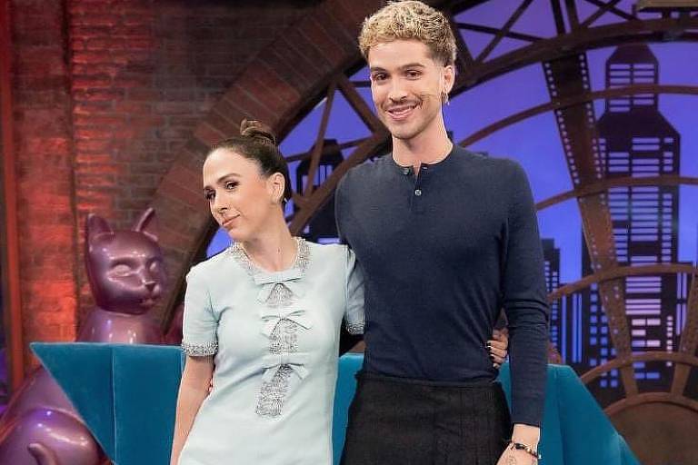 Na imagem, duas pessoas estão posando juntas em um cenário de programa de televisão. A mulher à esquerda usa um vestido azul claro com detalhes em renda e meias-calças pretas. O homem à direita veste uma camiseta preta e uma saia plissada escura, com um relógio visível em seu pulso. Ao fundo, há um sofá azul e uma decoração que inclui uma figura de gato roxo.