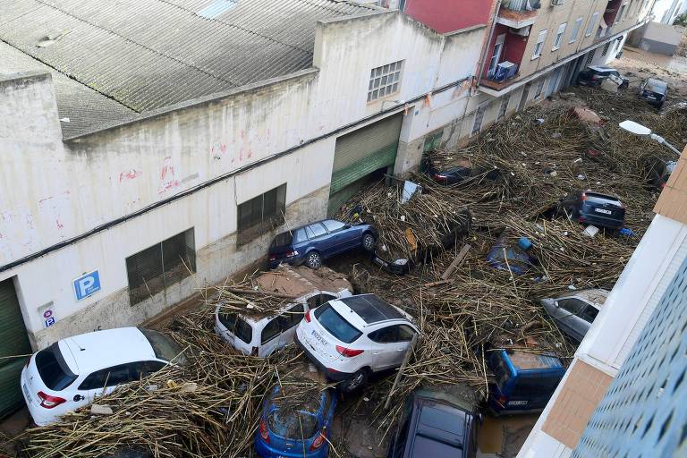 A imagem mostra uma rua urbana severamente afetada por um desastre natural, com vários carros cobertos por detritos, incluindo galhos e troncos. O cenário é de caos, com os veículos empilhados e obstruídos por materiais orgânicos, enquanto as paredes dos edifícios ao fundo estão visíveis. A área parece ter sido inundada, com água acumulada em algumas partes.