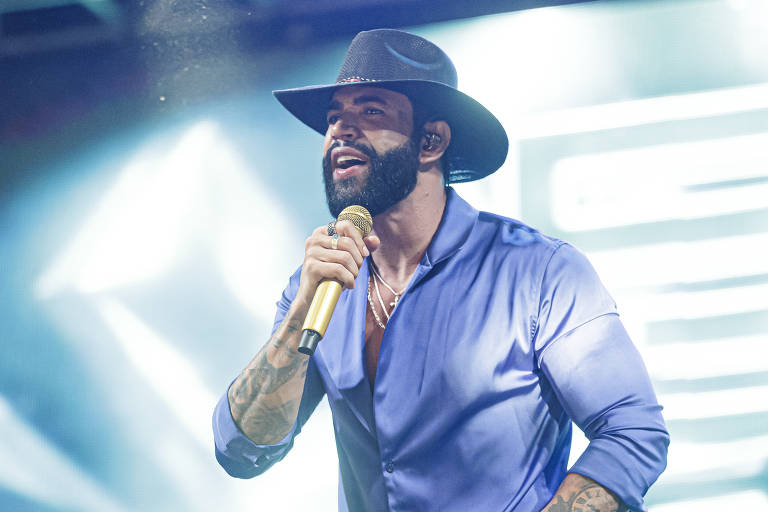 Um cantor está se apresentando em um palco iluminado， usando uma camisa azul e um chapéu preto. Ele segura um microfone dourado e parece estar cantando com intensidade. O fundo é iluminado com luzes brilhantes， criando um ambiente de show.