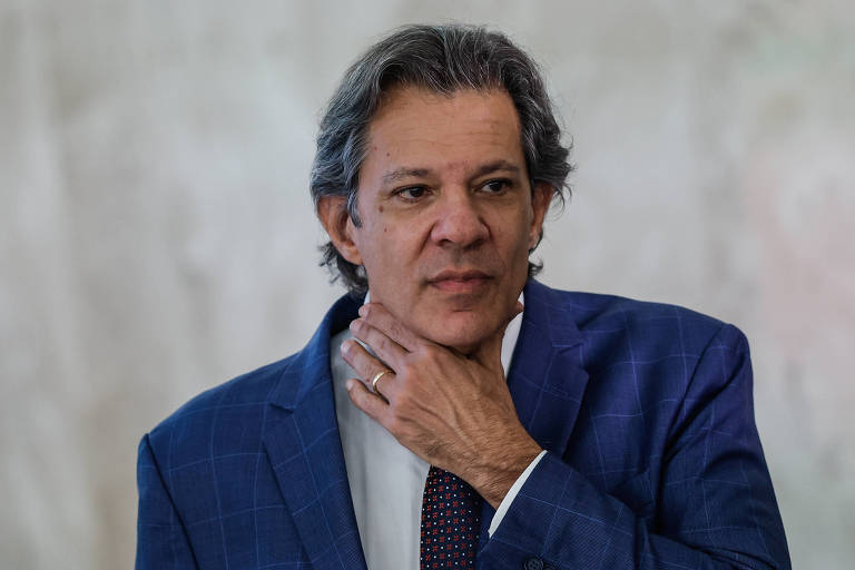 Um homem com cabelo grisalho e liso, vestindo um paletó azul e uma camisa branca com gravata. Ele está tocando o pescoço com a mão direita, aparentando estar pensativo. O fundo é desfocado, com uma tonalidade clara.
