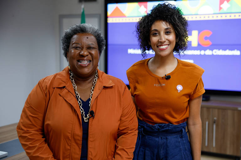 A imagem mostra duas mulheres sorrindo em um ambiente interno. A mulher à esquerda tem cabelo curto e grisalho, vestindo uma blusa azul e um casaco laranja, com um colar grande. A mulher à direita tem cabelo cacheado e usa uma blusa laranja e uma saia azul. Ao fundo, há uma tela com o texto 'MHC' e elementos coloridos.