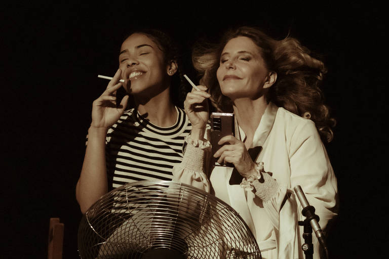 A imagem mostra duas mulheres em um ambiente escuro. A mulher à esquerda, com cabelo preso e usando uma camiseta listrada, está sorrindo enquanto segura um cigarro. A mulher à direita, com cabelo solto e ondulado, está com os olhos fechados, segurando um celular e também um cigarro. Entre elas, há um ventilador que parece estar ligado, com as lâminas visíveis. A iluminação é suave, criando um clima intimista.