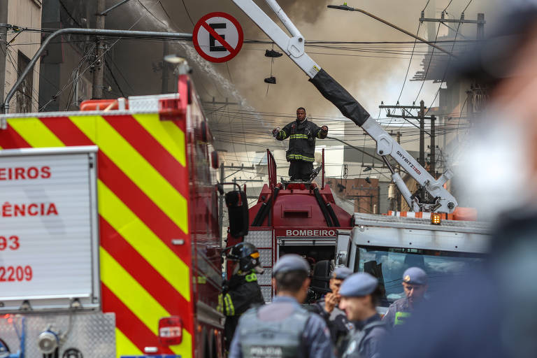 A imagem mostra uma cena de emergência com bombeiros e policiais em uma área urbana. Um caminhão de bombeiros vermelho com faixas amarelas está em primeiro plano, enquanto um bombeiro em uma escada mecânica está posicionado acima, aparentemente coordenando as operações. Ao fundo, há fumaça densa saindo de um edifício. Vários policiais estão visíveis, observando a situação.