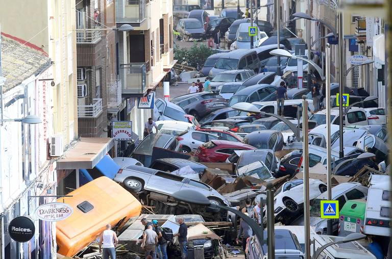 A imagem mostra uma rua urbana congestionada com muitos carros danificados e empilhados, resultado de um desastre, possivelmente uma inundação. Há pessoas tentando mover os veículos e a cena é marcada por desordem, com alguns carros virados e outros parcialmente submersos. Prédios residenciais estão visíveis ao fundo.