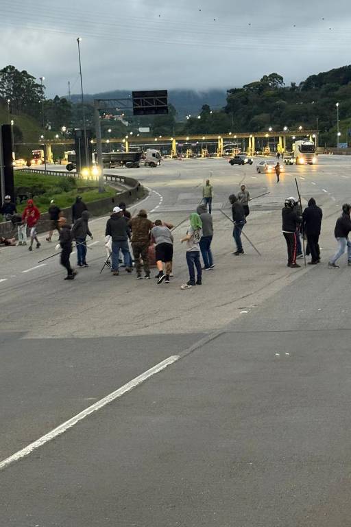 Um grupo de pessoas está reunido no meio de uma estrada. Algumas pessoas estão segurando varas ou bastões. Ao fundo, há uma praça de pedágio e veículos se aproximando. O céu está nublado e a atmosfera parece tensa.