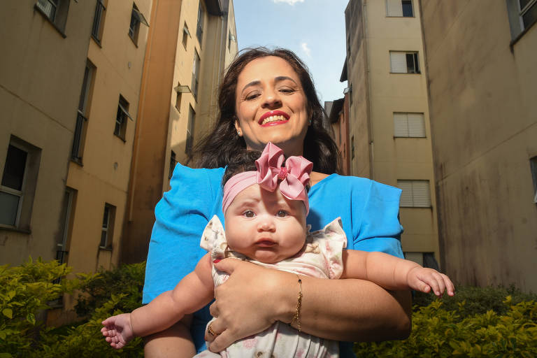 Uma mulher sorridente está segurando um bebê em seus braços. A mulher usa uma blusa azul e tem cabelo solto. O bebê, que usa um laço rosa na cabeça, parece estar olhando para a câmera. Ao fundo, há prédios de apartamentos e vegetação baixa na parte inferior da imagem, com um céu parcialmente nublado.