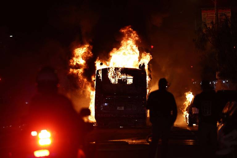 A imagem mostra um ônibus em chamas, com fogo e fumaça saindo de sua parte superior. A cena é noturna, com pessoas em silhueta observando o incêndio. Ao fundo, há luzes de veículos e uma atmosfera de caos.