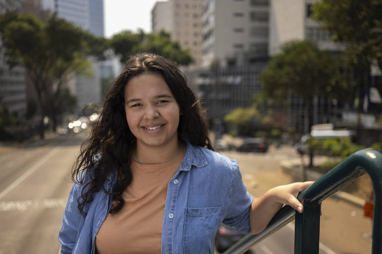 Uma jovem com cabelo cacheado e escuro sorri para a câmera enquanto se apoia em uma grade. Ela usa uma camiseta bege e uma camisa jeans. Ao fundo, há uma rua urbana com prédios e árvores, e alguns carros visíveis.