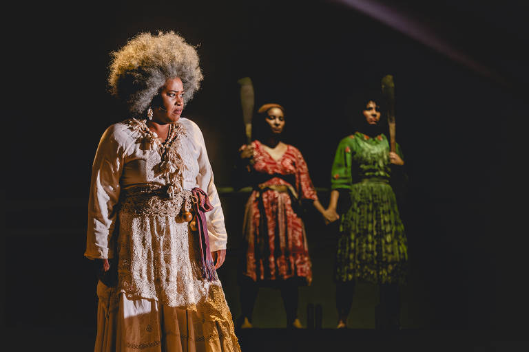 A imagem mostra três mulheres em uma cena de teatro. A mulher à frente tem cabelo volumoso e grisalho, vestindo uma blusa branca e uma saia longa com detalhes em renda. Ela parece estar em uma pose de destaque, com uma expressão séria. Atrás dela, duas mulheres estão de mãos dadas, uma vestindo um vestido vermelho e a outra um vestido verde. Ambas seguram bastões, e a iluminação destaca suas silhuetas contra um fundo escuro.