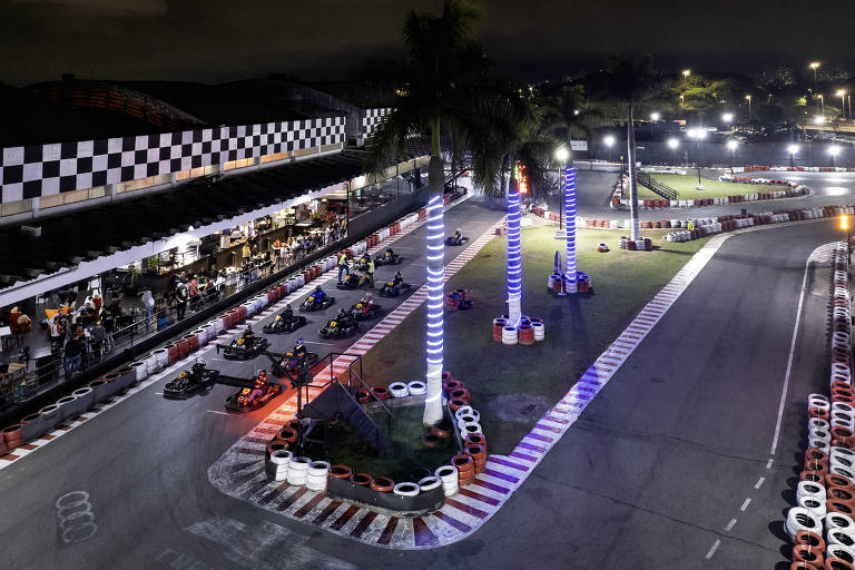 Imagem de uma pista de kart iluminada à noite, com karts alinhados na pista. Há palmeiras iluminadas e uma estrutura de arquibancada com bandeiras quadriculadas ao fundo. A pista é cercada por luzes brilhantes, criando um ambiente vibrante.