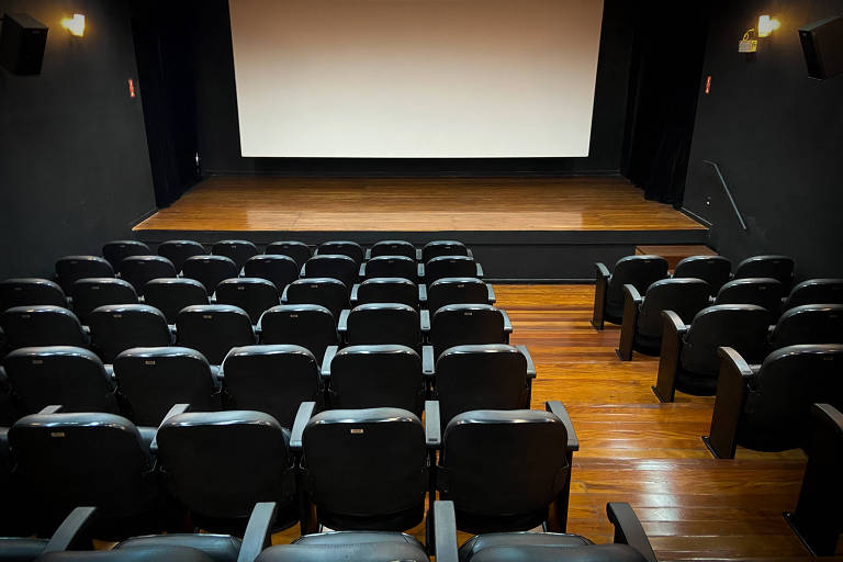 Imagem de um auditório vazio com cadeiras pretas dispostas em fileiras. No fundo, há um palco de madeira e uma tela grande e branca. As paredes são escuras, e a iluminação é suave, com algumas luzes nas laterais.