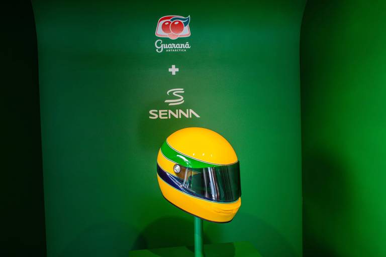 A imagem mostra um capacete de Fórmula 1 em um suporte, com as cores verde e amarelo, representando a bandeira do Brasil. O fundo é verde e apresenta os logotipos de 'Guaraná Antarctica Brands' e 'Senna' acima do capacete.