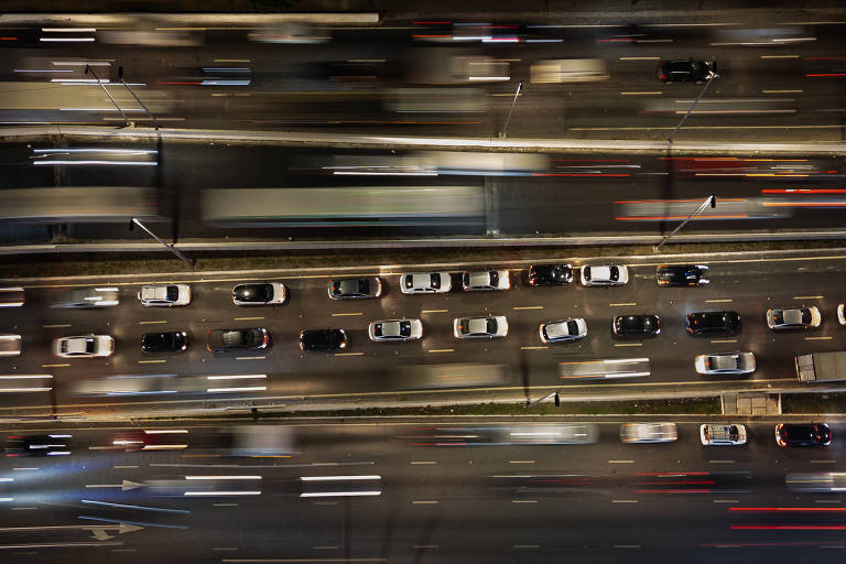 Imagem aérea de uma estrada movimentada à noite， mostrando uma fila de carros em movimento. Os veículos são predominantemente brancos， com algumas cores diferentes， e as luzes dos faróis e lanternas criam um efeito de movimento. A estrada é iluminada， e há um fluxo intenso de tráfego em ambas as direções.