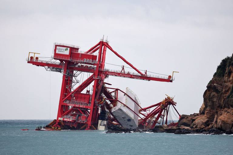 A imagem mostra um navio encalhado em uma estrutura de guindaste vermelho, inclinada em direção a uma rocha. O céu está nublado e o mar é calmo, com a estrutura do guindaste se destacando contra a paisagem rochosa ao fundo.