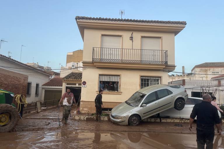 A imagem mostra uma casa de dois andares com uma fachada clara. Um carro está parcialmente em cima de uma plataforma ao lado da casa, enquanto o solo ao redor está coberto de lama. Algumas pessoas estão visíveis na cena, incluindo um homem com uma camisa clara e outro com uma camisa escura. Ao fundo, há um trator e outras estruturas. O céu está claro, indicando que a chuva pode ter cessado.