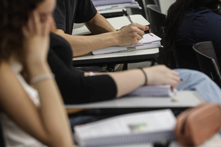 A imagem mostra um grupo de estudantes em uma sala de aula. Em primeiro plano， há duas mãos: uma segurando um caderno e a outra apoiada na mesa. Ao fundo， é possível ver mais estudantes， com alguns cadernos e canetas sobre as mesas. O ambiente parece ser de aprendizado， com foco nas atividades escolares.