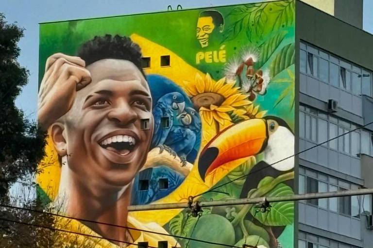 O mural retrata um jovem sorridente vestindo uma camisa amarela com detalhes verdes, simbolizando a seleção brasileira. Ao fundo, há elementos da natureza, como um tucano, flores e pássaros, além de uma imagem de Pelé. O mural é vibrante e colorido, refletindo a cultura brasileira.