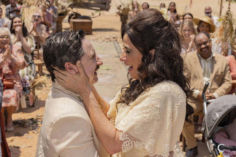 A imagem mostra um casal se olhando nos olhos, sorrindo e se tocando durante uma cerimônia de casamento ao ar livre. O homem usa um traje claro e tem um bigode, enquanto a mulher veste uma blusa de crochê clara e tem cabelos longos e soltos. Ao fundo, há uma plateia de convidados aplaudindo e sorrindo, com um cenário de terra e vegetação ao redor.