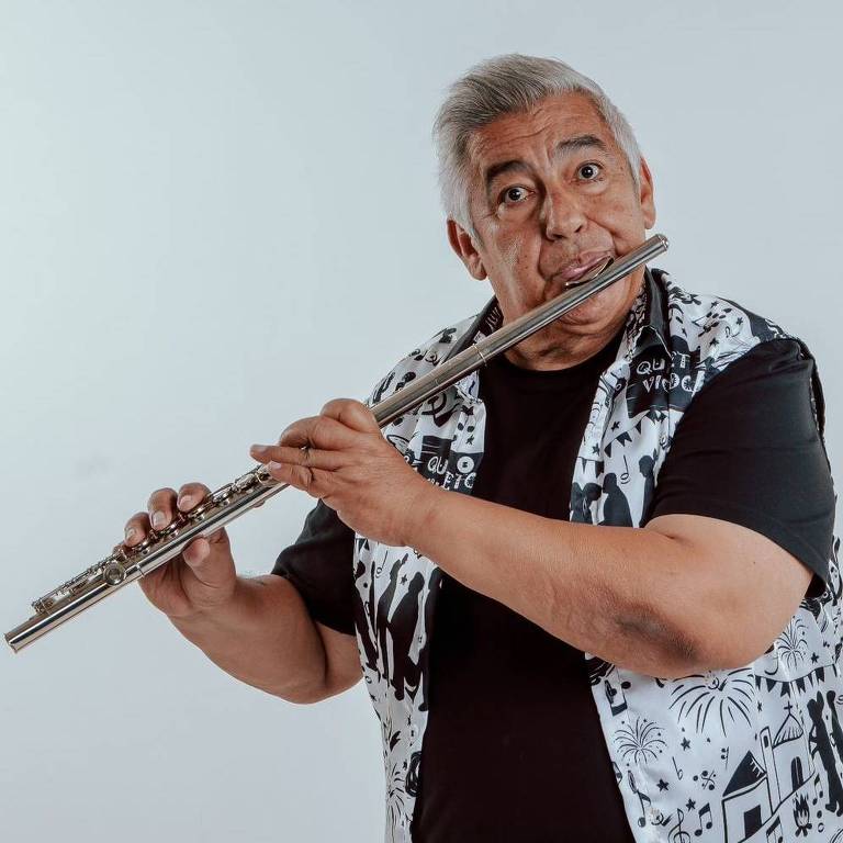 Um homem de cabelos grisalhos está tocando uma flauta. Ele usa uma camiseta preta e uma camisa de estampa branca e preta. O fundo é claro e neutro.