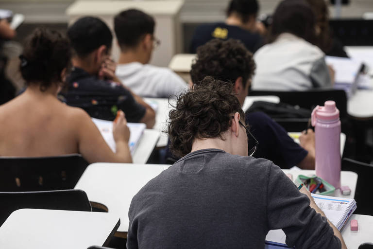 A imagem mostra um grupo de estudantes em uma sala de aula， vistos de costas. Alguns alunos estão concentrados em suas atividades， escrevendo em cadernos ou livros. Um estudante à frente tem uma garrafinha rosa ao seu lado. O ambiente parece ser de uma instituição de ensino， com mesas e cadeiras organizadas.