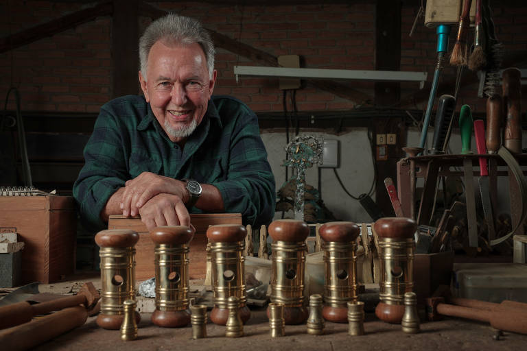 Um homem sorridente está posicionado em um ateliê de marcenaria, apoiando-se em uma mesa de trabalho. Ele usa uma camisa xadrez verde e tem cabelo grisalho. À sua frente, há uma linha de objetos de metal dourado, possivelmente peças de marcenaria, e várias ferramentas estão visíveis ao fundo, junto a caixas de madeira e outros materiais de trabalho.