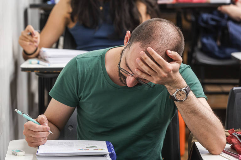 Um homem com cabelo ralo, usando uma camiseta verde, está sentado em uma mesa de aula. Ele tem a mão na cabeça, parecendo estressado ou preocupado, enquanto escreve em um caderno. Ao fundo, é possível ver uma mulher com cabelo longo e escuro, também em uma mesa de aula.