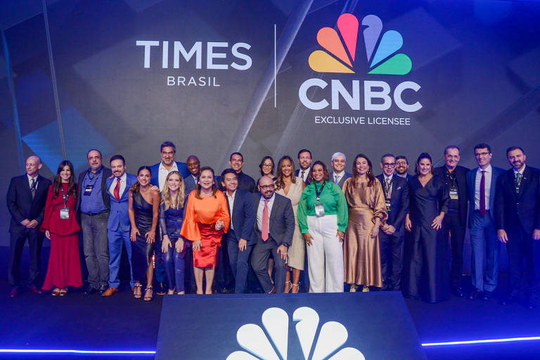 A imagem mostra um grupo de pessoas posando em um evento, com o logotipo da Times Brasil e da CNBC ao fundo. As pessoas estão vestidas de forma variada, com algumas em trajes formais e outras em roupas mais casuais. O ambiente é bem iluminado e decorado, com um palco ao fundo. O grupo parece estar celebrando ou participando de uma conferência.