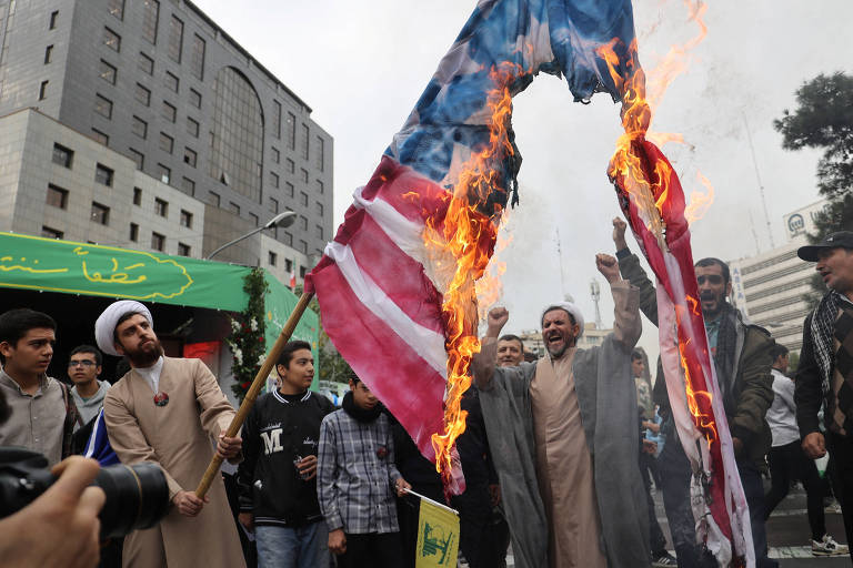 A imagem mostra um grupo de pessoas em um protesto, com uma bandeira americana em chamas. Algumas pessoas estão levantando as mãos em sinal de celebração ou protesto, enquanto outras observam. O ambiente é urbano, com edifícios ao fundo.