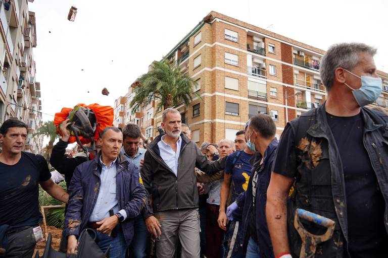 A imagem mostra um grupo de pessoas em uma área urbana, onde um homem de cabelo grisalho, vestido com um casaco cinza, é cercado por várias pessoas. Algumas delas estão usando máscaras faciais. Ao fundo, há um prédio de apartamentos e árvores. Um dos homens carrega um objeto laranja, possivelmente uma mochila ou equipamento de resgate. O ambiente parece estar em recuperação após um evento adverso.