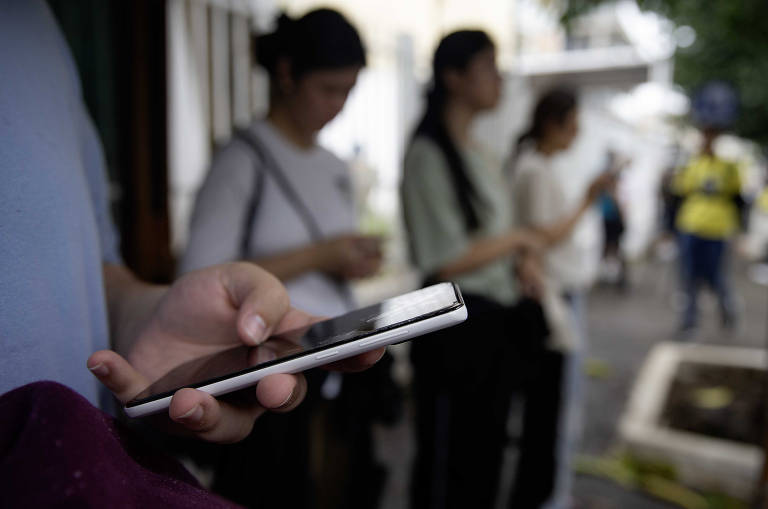 A imagem mostra um grupo de pessoas em pé, aparentemente esperando, enquanto usam seus smartphones. Em primeiro plano, uma mão segura um smartphone. Ao fundo, outras pessoas estão visíveis, algumas com os olhos voltados para seus dispositivos móveis.