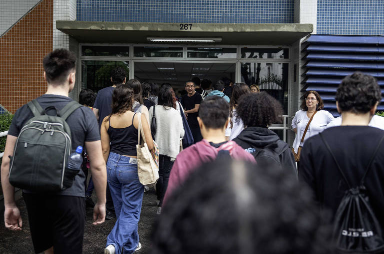 A imagem mostra um grupo de pessoas caminhando em direção à entrada de um edifício. Algumas pessoas estão de costas, enquanto outras estão de frente. O ambiente parece ser urbano, com uma fachada colorida ao fundo. A maioria das pessoas está vestindo roupas casuais e algumas carregam mochilas. Há uma pessoa em destaque na entrada, possivelmente interagindo com o grupo.