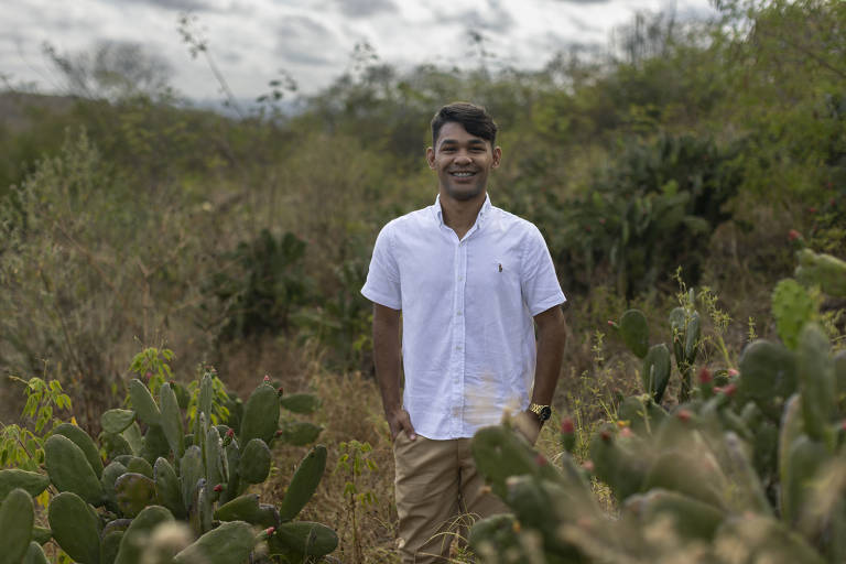 Um jovem está posando em um campo aberto, cercado por vegetação e cactos. Ele usa uma camisa branca de botão e calças claras, com as mãos nos bolsos. O fundo é composto por plantas secas e cactos, sob um céu nublado.