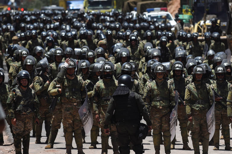 A imagem mostra uma grande formação de soldados em um ambiente ao ar livre. Os soldados estão vestidos com uniformes camuflados e usam capacetes, alguns deles segurando escudos. A formação é densa, com muitos soldados alinhados em várias fileiras. Ao fundo, é possível ver veículos e uma área de construção.